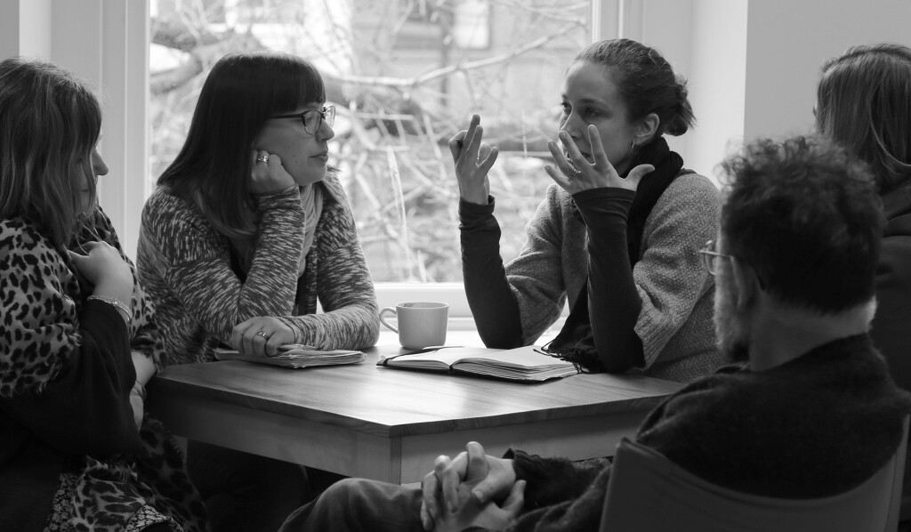 Black and white photo of Katharine sitting at a table, in conversation with a group of other people.