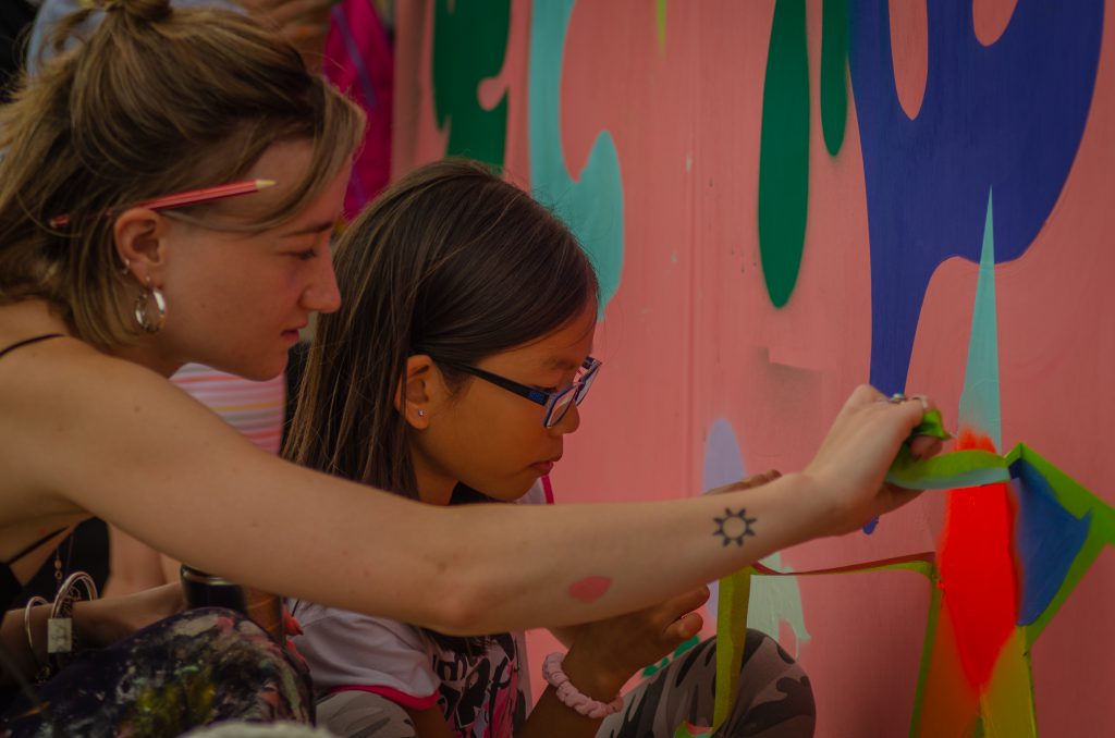 A young participant and a practitioner work on a large outdoor mural at the Wallacetown fun day.