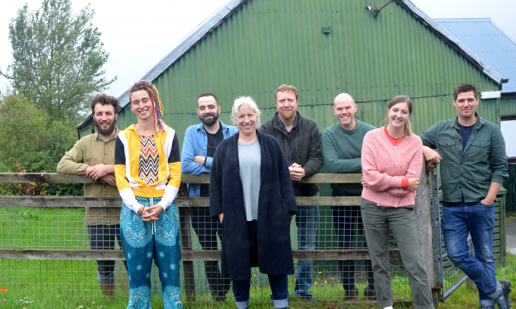 Eight people stand smiling at the camera, in front of a green, corrugated iron shed.