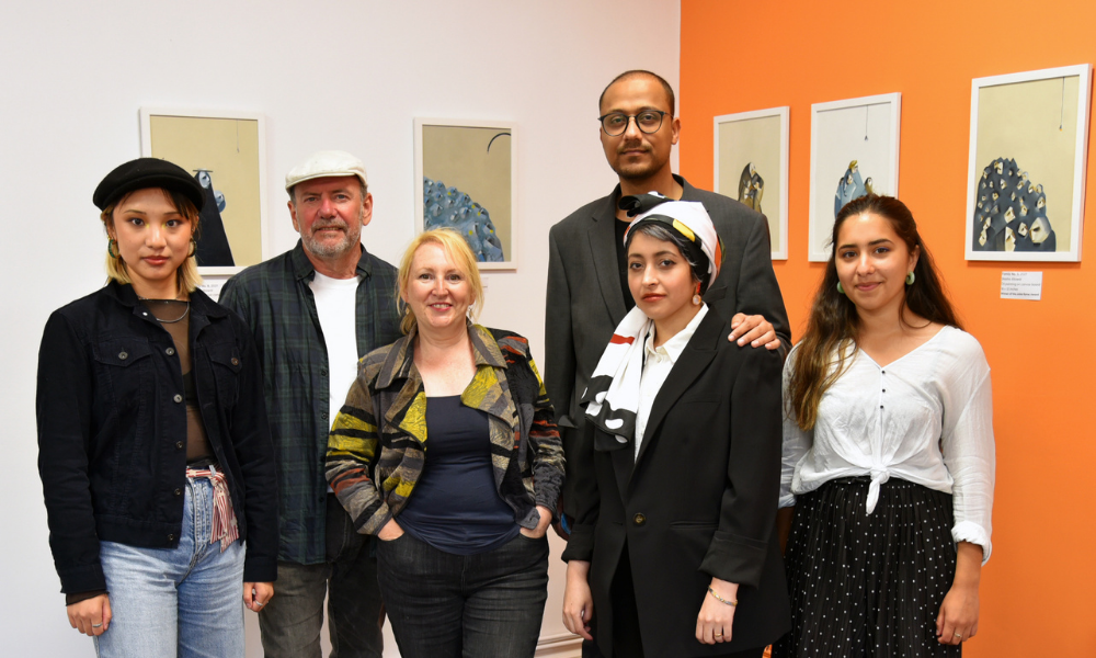 Six people, the artists and coordinators for Art27@Southside, stand smiling at the camera in an orange and white gallery space, with paintings on the walls.