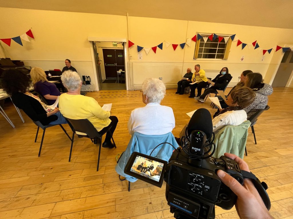 A group of people sit in a semi circle in what looks like a town hall with a wooden floor. We can see they are being filmed, because the photo has been taken from just behind the video camera.