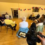 A group of people sit in a semi circle in what looks like a town hall with a wooden floor. We can see they are being filmed, because the photo has been taken from just behind the video camera.