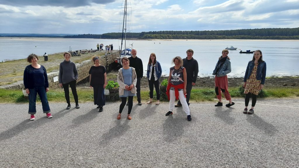 10 people stand by a pier in the sunshine, with Findhorn Bay spread out behind them.