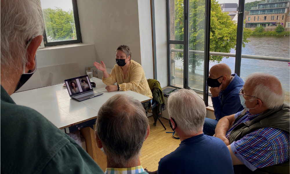 An artist in a yellow short and black mask sits at a desk, gesticulating to a laptop. Around her, a group of five people listen intently.