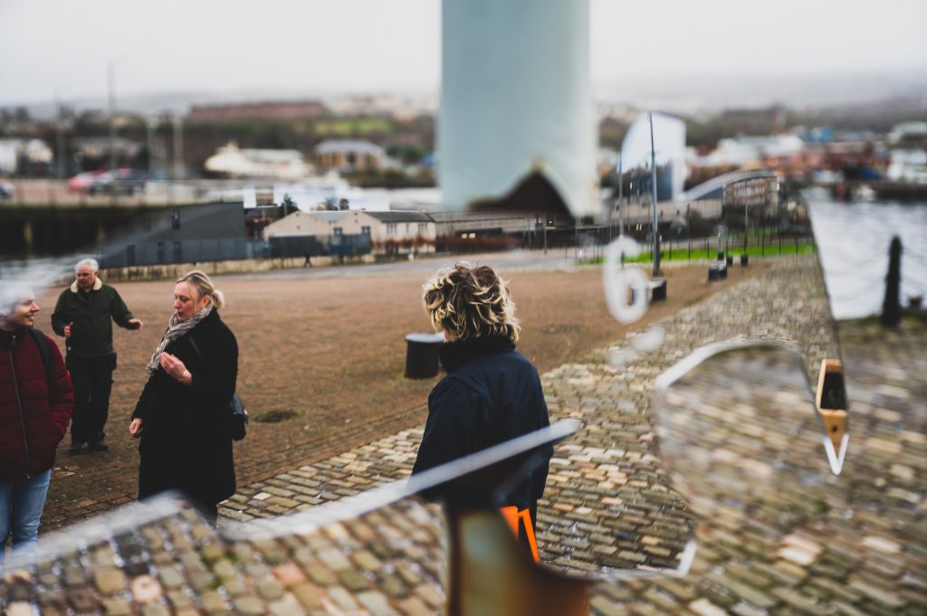 Framing the image is an outline of a bird, which holds a reflection of the dockside of Greenock Inverclyde, there is a faint number 6 carved in the centre, in the background stand three people facing different directions