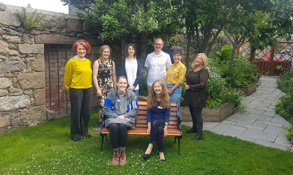 A group of eight people, the coordinators of Safe Harbour: Open Sea, stand smiling at the camera in a lush, green garden.