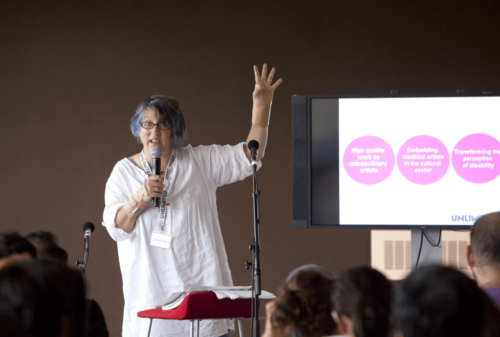 Jo stands as the speaker at an event. She wears a long white top and has her left arm raised in the air. She holds a microphone in her right hand.