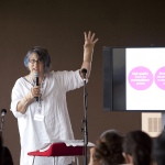 Jo stands as the speaker at an event. She wears a long white top and has her left arm raised in the air. She holds a microphone in her right hand.