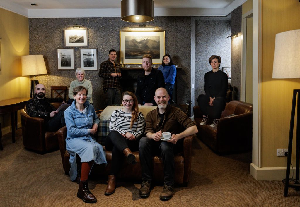 Nine people sit or stand in a room, gathered around a sofa. There are lamps and pictures in the background.