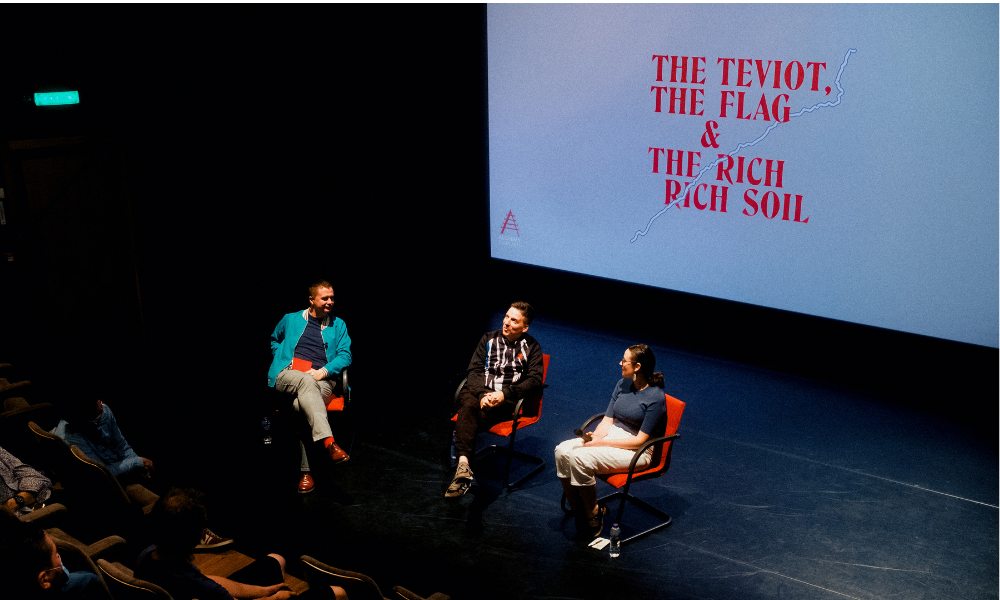 Three people sit in bright red chairs on a darkened stage, speaking to the gathered audience. Behind them, the logo for The Teviot, The Flag & The Rich Rich Soil is projected on a large cinema screen.