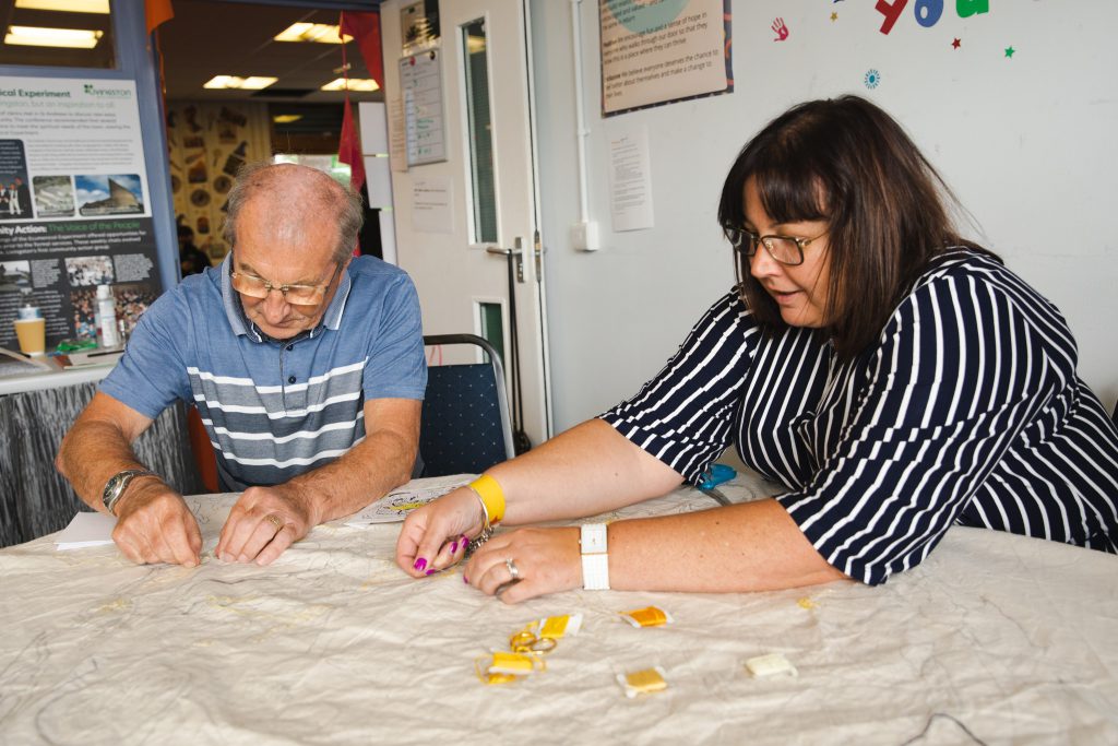 Two people sit at a table, which is covered by tapestry fabric. Their hands are busy stitching, and they are focussed intently on the task.