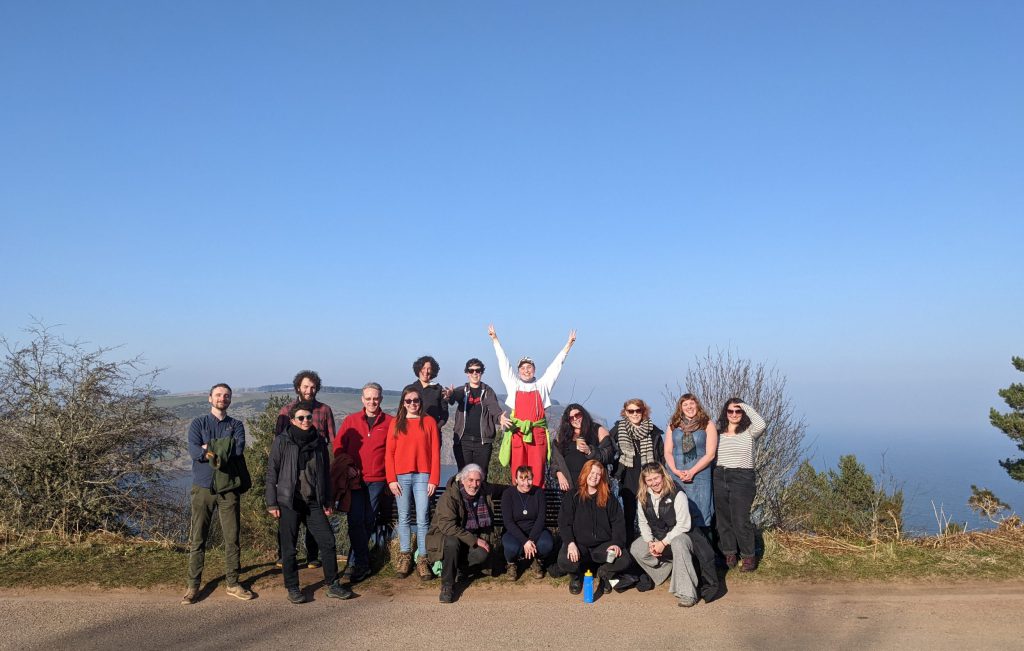 A group of people pose on top of a hill. There is a beautiful blue sky. Everyone is smiling and the person in the middle has their arms high in the air.