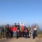 A group of people pose on top of a hill. There is a beautiful blue sky. Everyone is smiling and the person in the middle has their arms high in the air.