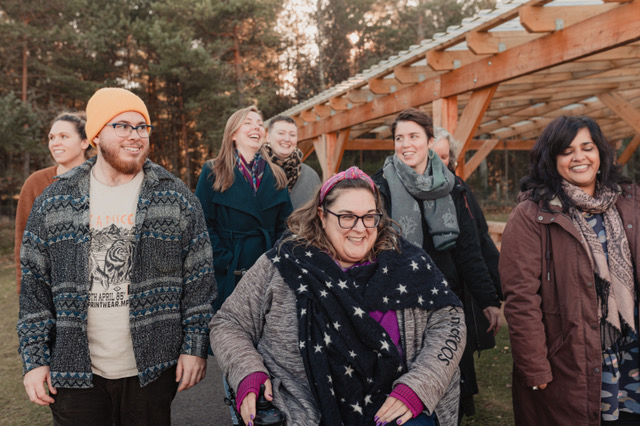 A group of seven people walk toward the camera. They are smiling and laughing together outdoors on a lovely autumnal day.