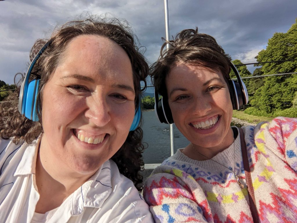 Kathryn Welch and Natalia Palombo grin at the camera, wearing big, blue ear muffs. They are on a bridge overlooking the River Spey in Aberlour.