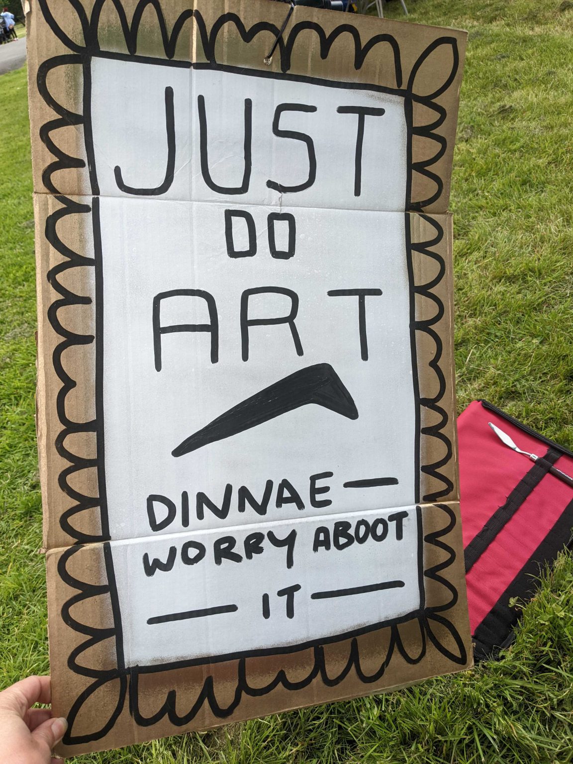 A brown cardboard sign with the words "Just do art, dinnae worry about it" and an upside-down nike-style tick.