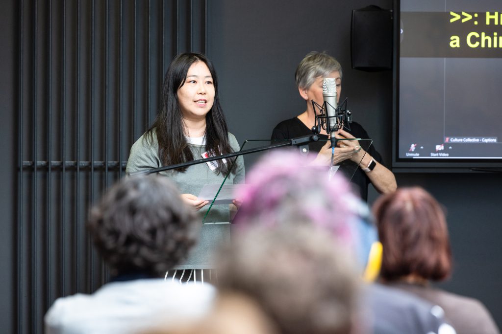 Elaine stands at a lectern and microphone at the front of a room full of people. A BSL interpreter stands to her side.