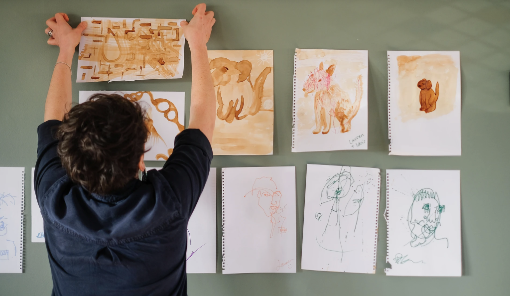 A person stands with their back to the camera, attaching a series of drawings to a green wall.